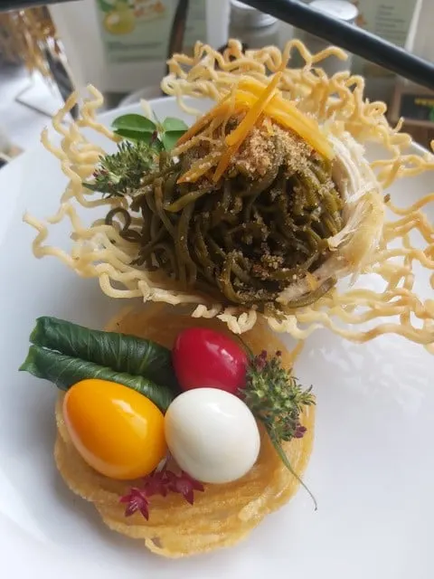 Mi hijau disajikan di atas sarang mi goreng dengan telur rebus dan sayuran di piring putih