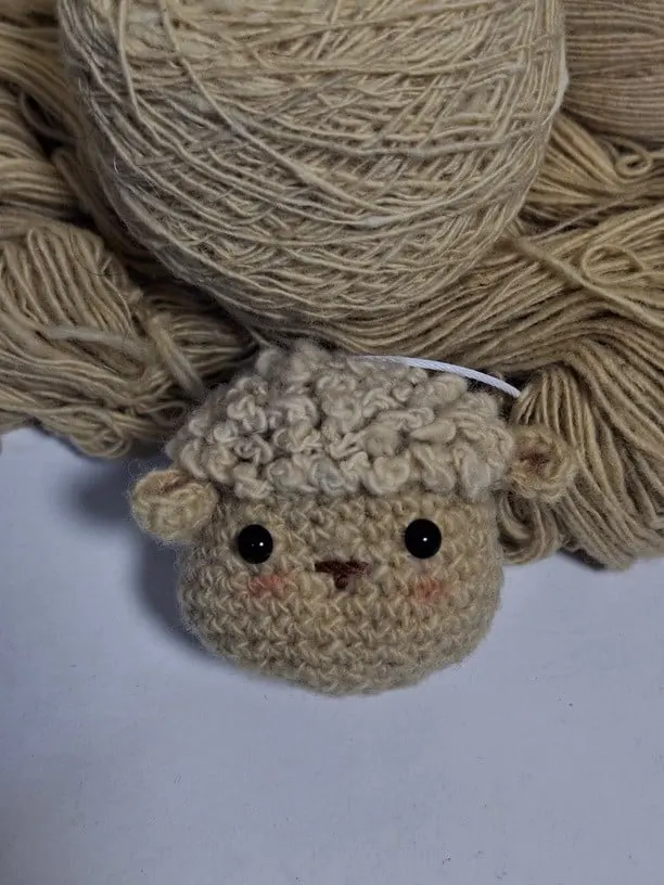 Boneka rajut domba krem bermata hitam dengan bulu keriting di depan gulungan benang krem