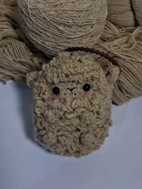 Boneka rajut domba krem berbulu keriting dengan tali gantungan cokelat di depan gulungan benang