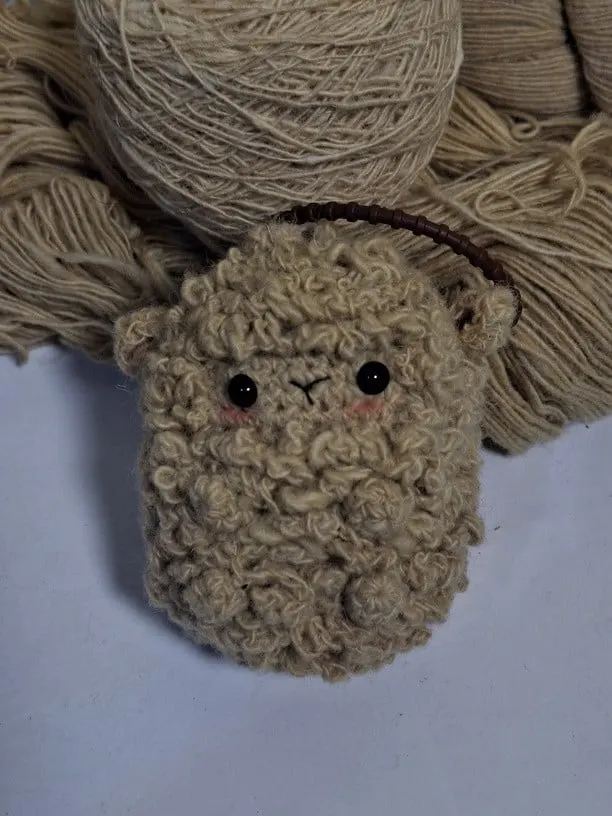 Boneka rajut domba krem berbulu keriting dengan tali gantungan cokelat di depan gulungan benang