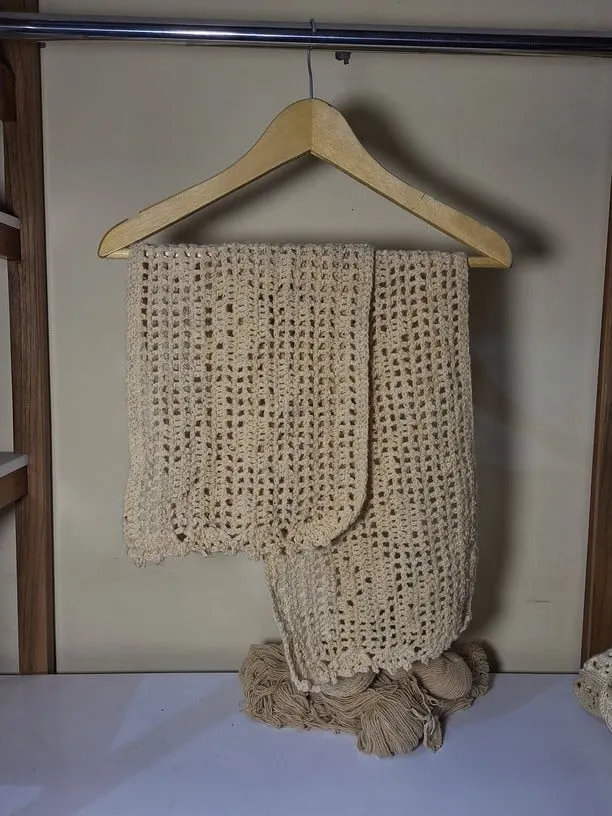 Syal rajut krem bermotif jaring terlipat pada hanger kayu di atas gulungan benang