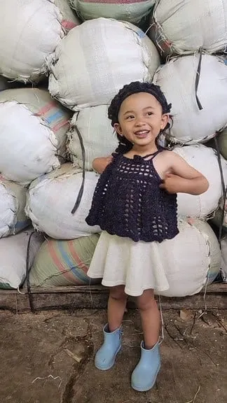 Anak perempuan memakai atasan rajut biru tua dan bando rajut di depan tumpukan karung putih