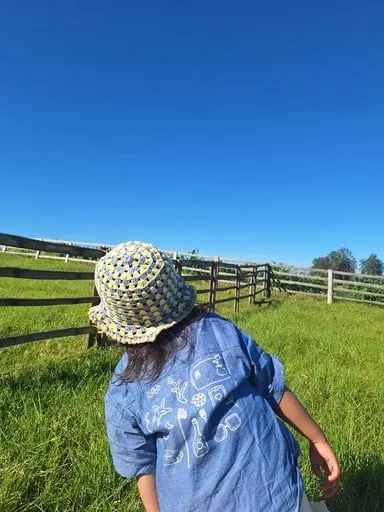 Anak berkemeja denim memakai topi bucket rajut biru dan krem di padang rumput berpagar kayu
