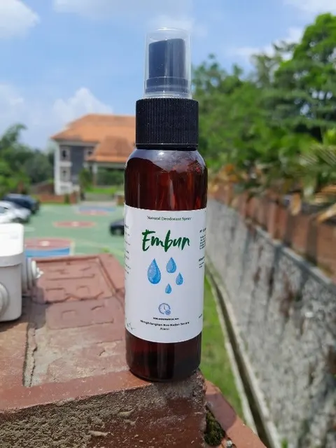 Botol spray cokelat berlabel putih Embun Natural Deodorant Spray di atas pagar bata, latar langit biru