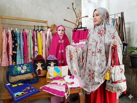 Perempuan memakai mukena abu motif mawar sambil membawa tas kain, di samping mukena anak, tas dan rak baju anak