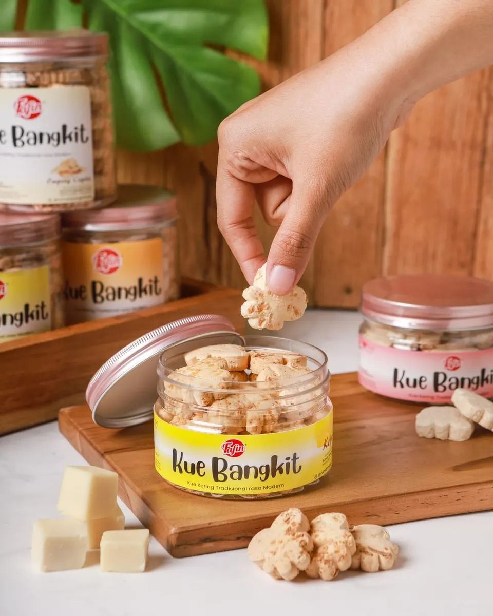 Tangan mengambil kue bangkit dari toples berlabel kuning Fafin, dengan toples lain dan potongan keju di sekitarnya