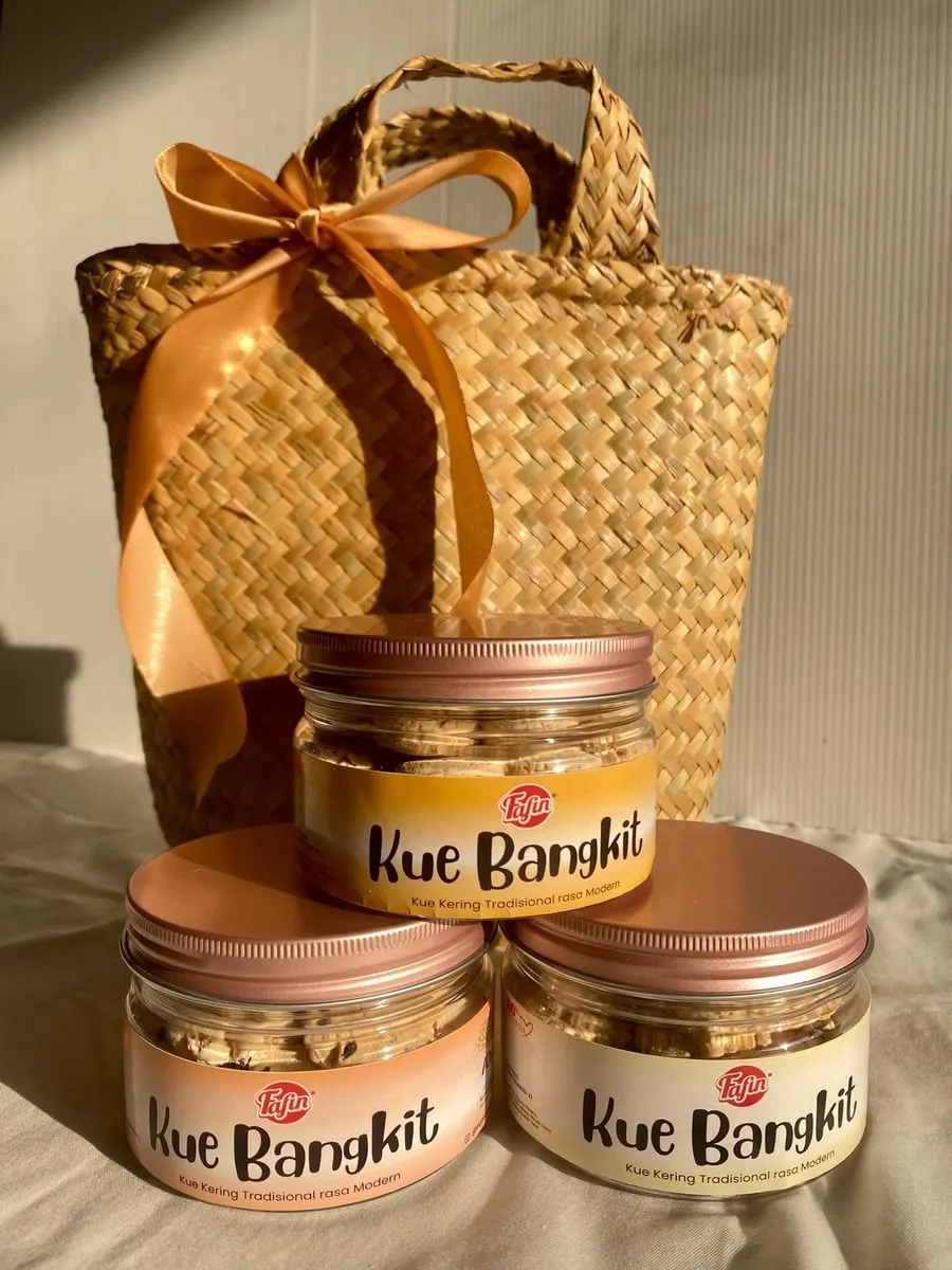Tiga toples Kue Bangkit Fafin bertutup warna emas merah muda di depan keranjang anyaman berpita oranye
