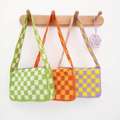 Tiga tas selempang rajut motif kotak-kotak hijau, oranye dan ungu kuning tergantung di gantungan kayu