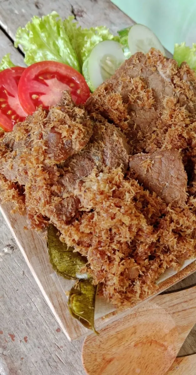 Potongan dendeng ragi sapi berbalut parutan kelapa goreng di atas papan kayu dengan selada, tomat dan timun