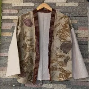 Outer ecoprint motif daun cokelat dan hijau berlist lurik, tergantung di depan dinding batu