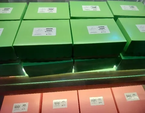 Tumpukan kotak kue hijau dan merah muda berlabel putih di rak berlampu