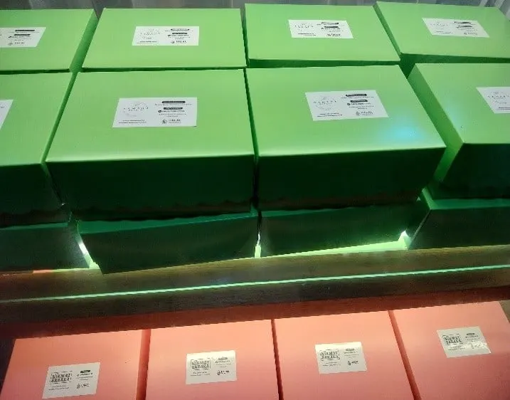 Tumpukan kotak kue hijau dan merah muda berlabel putih di rak berlampu