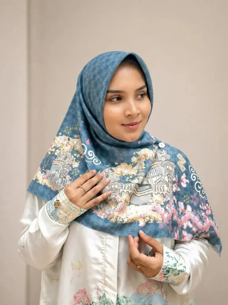 Model memakai hijab segi empat biru bermotif ilustrasi bangunan dan bunga dengan atasan satin putih