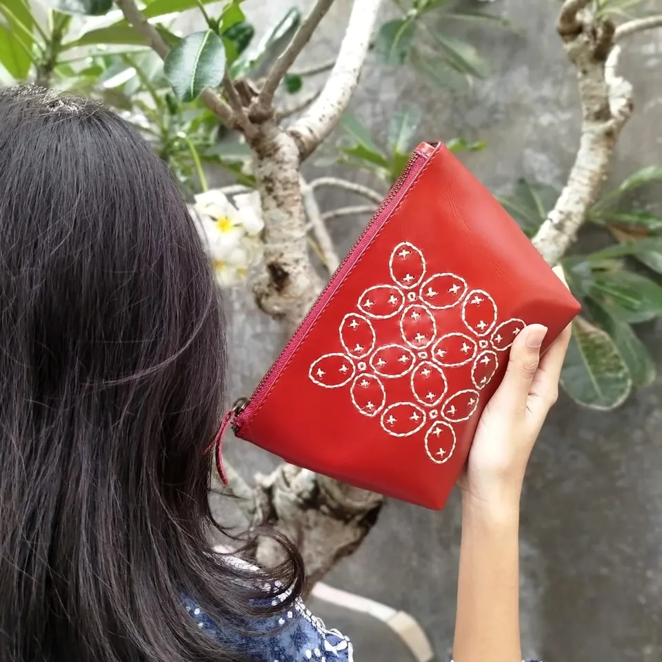 Perempuan berambut panjang mengangkat pouch kulit merah berjahit motif putih di dekat pohon