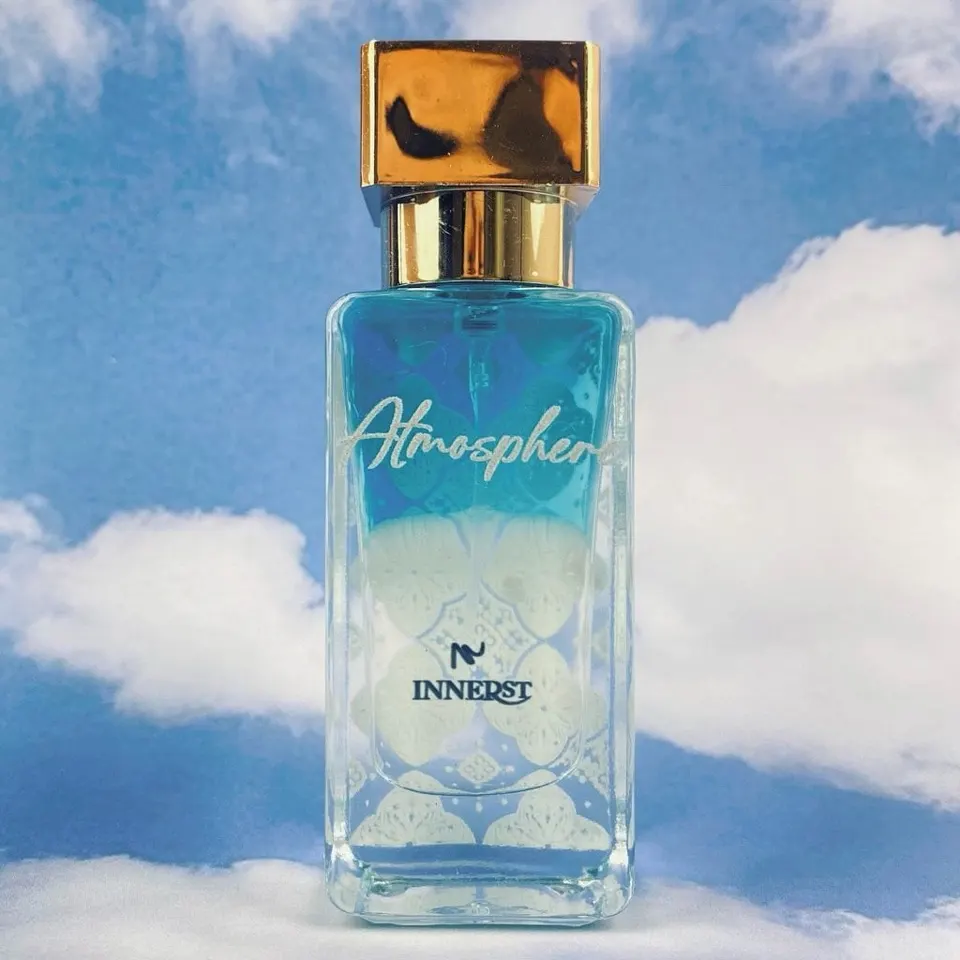 Botol parfum kaca tinggi Innerst Atmosphere bergradasi biru dan putih bertutup emas, berlatar langit berawan