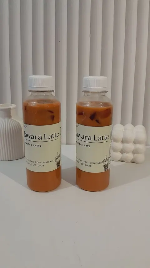 Dua botol thai tea oranye berlabel Jawara Latte Thai Tea Latte berdiri di meja putih di antara hiasan putih