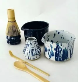 Mangkuk keramik bercerat biru tua dan putih bercorak biru, pengocok matcha bambu, dan sendok bambu di latar putih