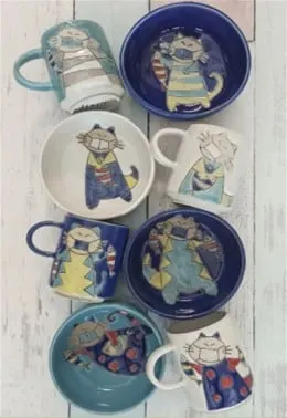 Mug dan mangkuk keramik bergambar kucing bermasker berwarna biru, putih, dan hijau tosca di atas meja kayu putih