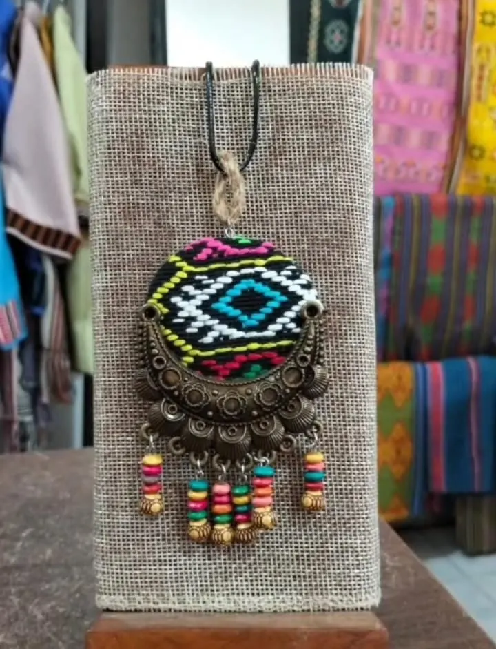 Kalung liontin bulat berhias tenun hitam, putih dan biru dengan bingkai logam dan juntaian manik kayu