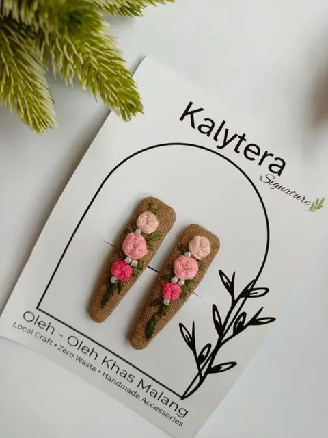 Sepasang jepit rambut berbalut kain cokelat dengan hiasan mawar sulam merah muda pada kartu Kalytera Signature
