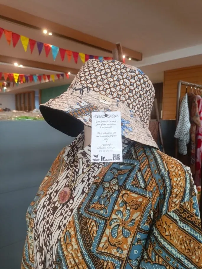 Bucket hat batik cokelat dengan label gantung Kalytera pada manekin berkemeja batik di sebuah toko