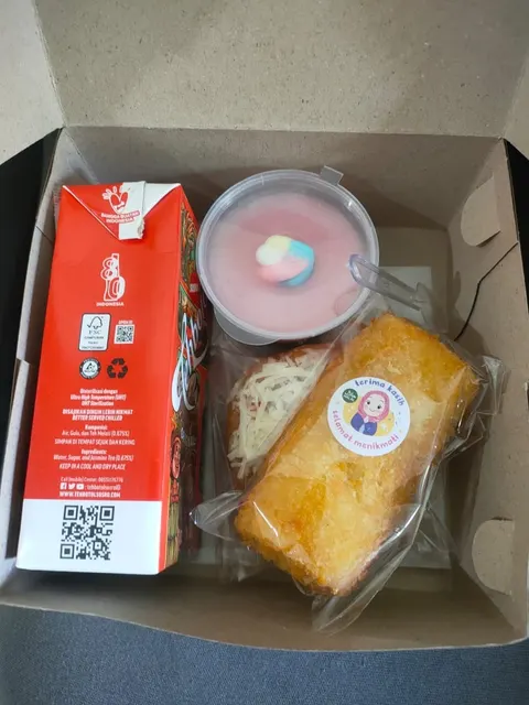 Kotak snack berisi minuman kotak merah, puding cup merah muda, kue dan roti berstiker dalam plastik