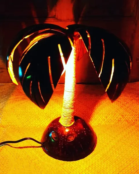 Lampu hias berbentuk pohon kelapa dari batok kelapa dengan batang berlilit tali, menyala jingga di atas tikar