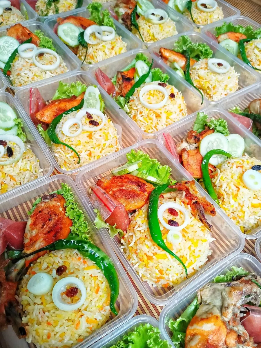 Kotak plastik berisi nasi basmati kuning, ayam panggang, cabai hijau, irisan bawang, mentimun dan kismis