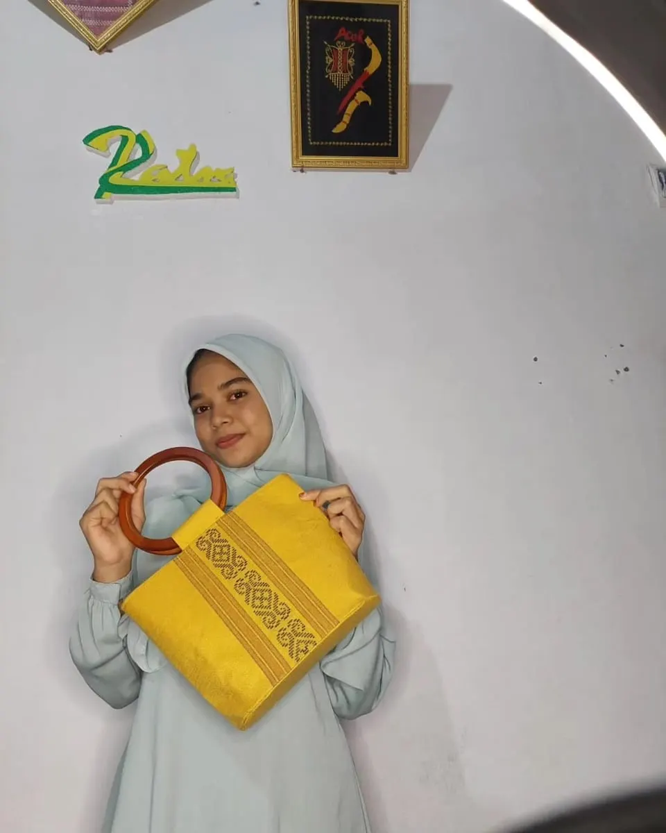 Perempuan berhijab memegang tas tangan kuning bergagang kayu bulat dengan pita bordir motif Aceh