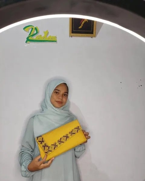 Perempuan berhijab memegang clutch kuning berbentuk amplop dengan bordir motif Aceh hitam di tepi tutupnya