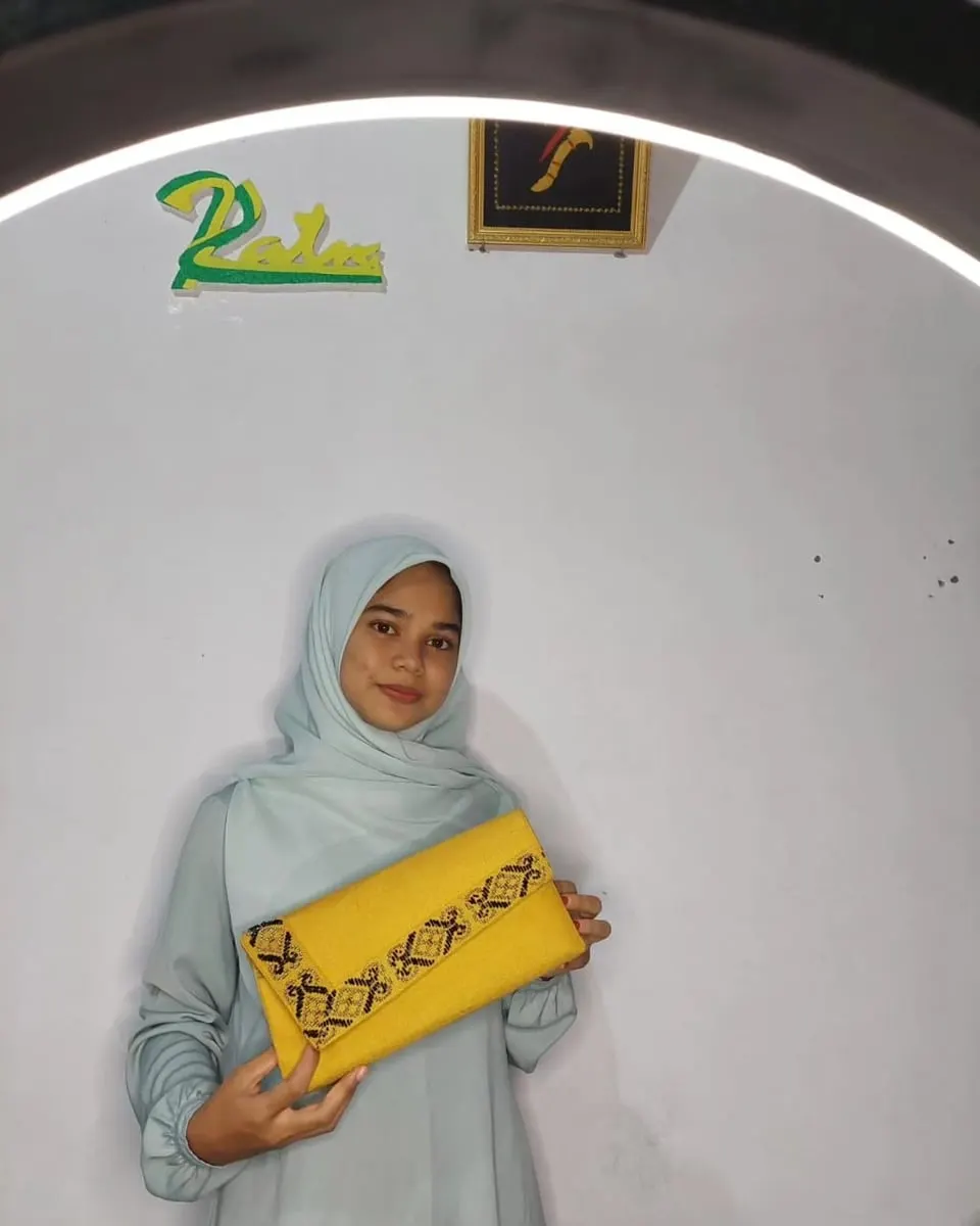 Perempuan berhijab memegang clutch kuning berbentuk amplop dengan bordir motif Aceh hitam di tepi tutupnya