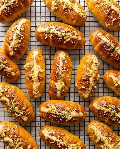 Deretan roti lonjong keemasan dengan topping saus krim dan bawang putih di atas rak kawat