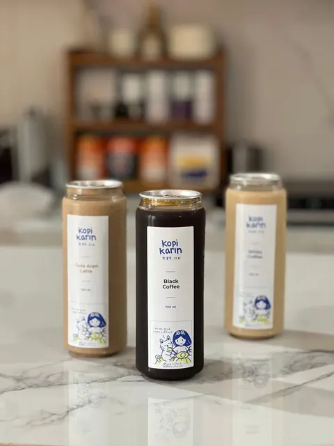 Tiga kaleng bening Kopi Karin varian Gula Aren Latte, Black Coffee dan White Coffee di atas meja marmer