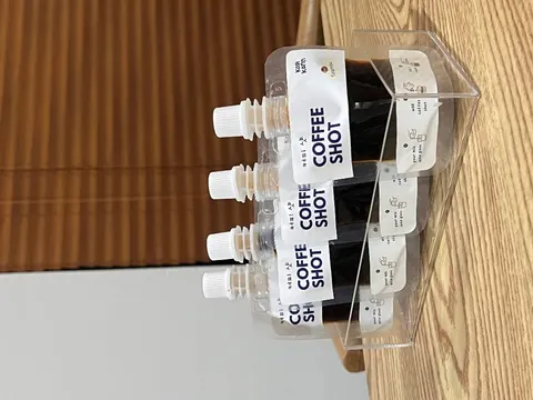 Empat kemasan pouch Coffee Shot berisi kopi hitam tersusun di rak akrilik yang menempel pada papan kayu