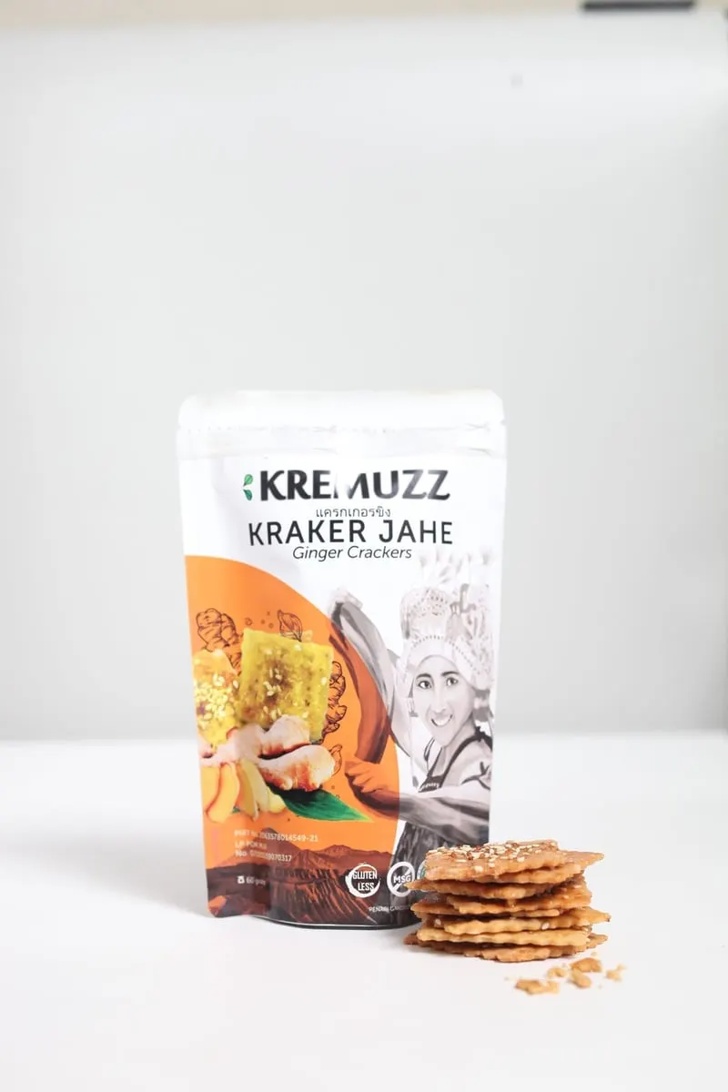 Kemasan pouch Kremuzz Kraker Jahe oranye putih dengan tumpukan kraker bertabur wijen di sampingnya