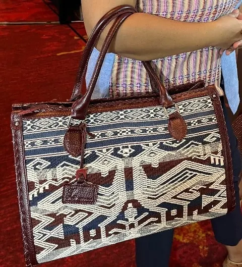 Tangan menjinjing tas kotak bermotif tenun biru, putih dan cokelat dengan pegangan dan list kulit cokelat