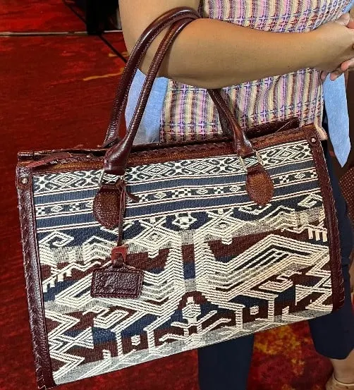 Tangan menjinjing tas kotak bermotif tenun biru, putih dan cokelat dengan pegangan dan list kulit cokelat