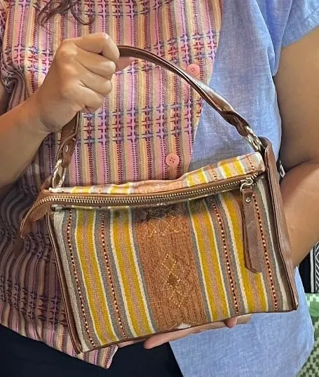 Tangan memegang tas kecil tenun bergaris kuning, cokelat dan merah muda dengan tali kulit cokelat