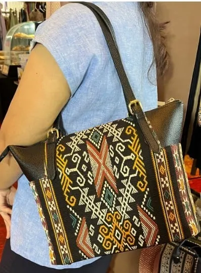 Perempuan menyandang tas bahu tenun hitam bermotif kuning dan merah dengan tali kulit hitam