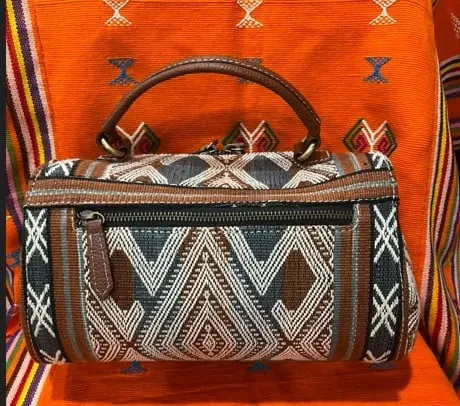 Tas tabung tenun motif belah ketupat biru putih dengan pegangan kulit cokelat di atas kain oranye
