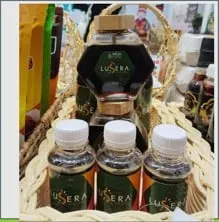 Jar madu segi enam dan tiga botol plastik kecil madu Lusera berlabel hijau di dalam keranjang rotan