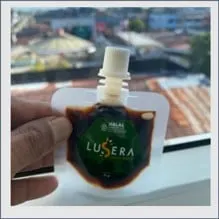 Tangan memegang pouch bercorong berisi madu gelap berlabel hijau Lusera dengan logo halal, berlatar atap rumah
