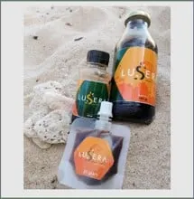 Dua botol dan satu pouch madu Lusera berlabel oranye dan hijau di atas pasir dekat batu karang