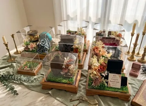 Deretan kotak hantaran akrilik beralas kayu berisi seserahan dengan hiasan bunga dan lilin di atas meja