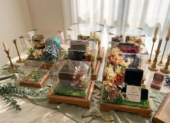 Deretan kotak hantaran akrilik beralas kayu berisi seserahan dengan hiasan bunga dan lilin di atas meja