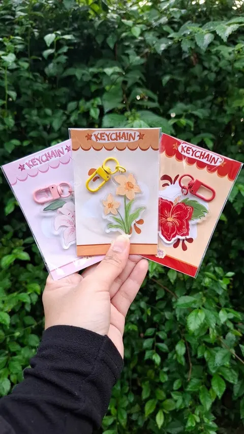 Tangan memegang tiga gantungan kunci akrilik bergambar bunga lili, narsis dan kembang sepatu pada kartu Keychain