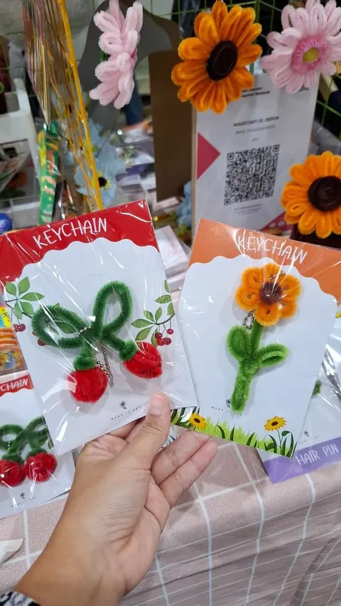 Tangan memegang gantungan kunci bulu kawat berbentuk ceri dan bunga matahari pada kartu Keychain di stan bazar