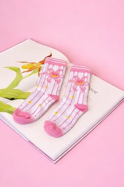 Sepasang kaus kaki putih bergaris merah muda bergambar bunga di atas buku terbuka berlatar merah muda