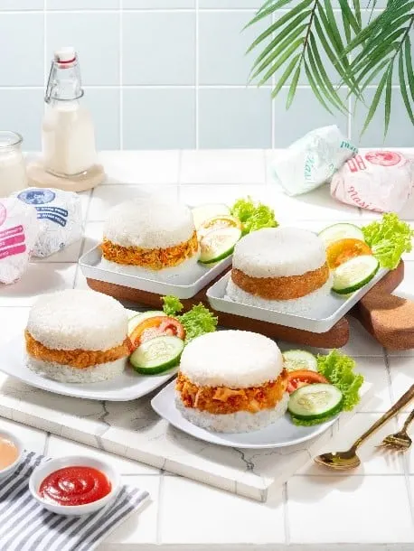 Empat nasi kepal isi ayam di piring putih dengan irisan timun, selada dan saus tomat di atas meja keramik
