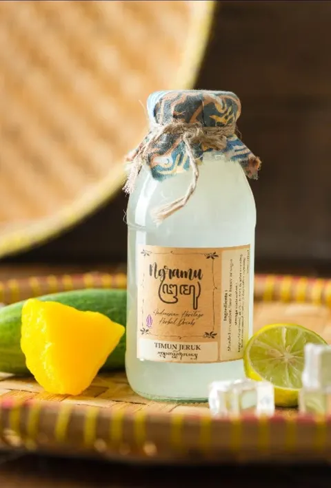Botol kaca Timun Jeruk berlabel Ngramu bertutup kain batik biru di nampan anyaman, dengan timun dan jeruk nipis